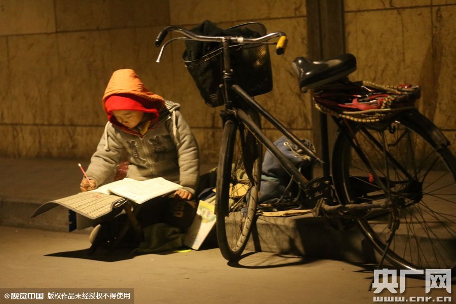 鄭州中原路京廣鐵路大橋下涵洞的馬路牙子邊，一位戴著紅色辣椒帽的小男孩趴在一個(gè)小馬扎上，上面鋪著一張車墊，在微弱的燈光下寫作業(yè)