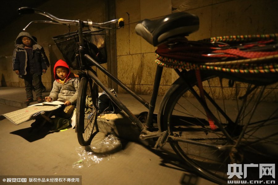 2015年11月8日晚，鄭州，7歲男孩涵洞橋下陪父母練攤，路邊燈下坐小馬扎上寫作業(yè)。