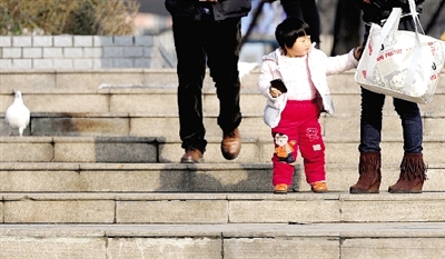 昨天中午，一女孩拉著家長的手在紫荊山公園曬太陽，氣溫升高，孩子的棉衣解開了扣子。